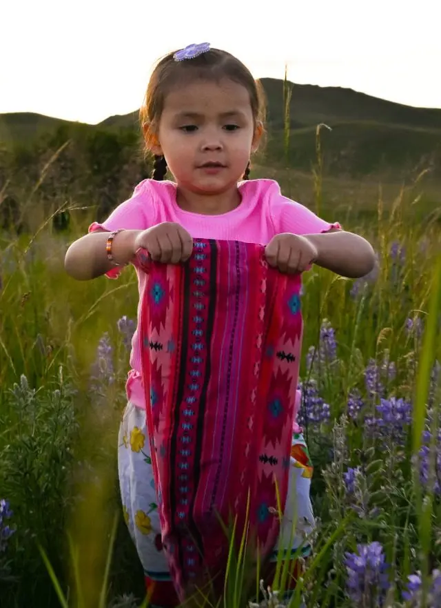 A young girl in a pink shirt holds up a patterned fabric in a field of wildflowers.