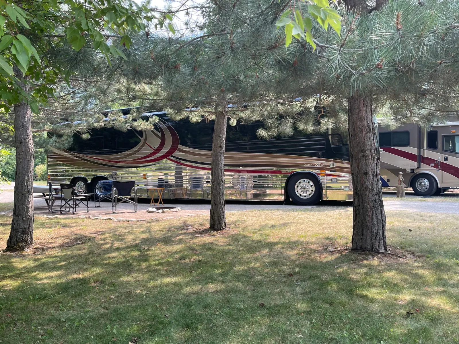 A luxury Prevost motorcoach with its awning extended and camp chairs set up on a shaded pull-through site at Campground St. Regis