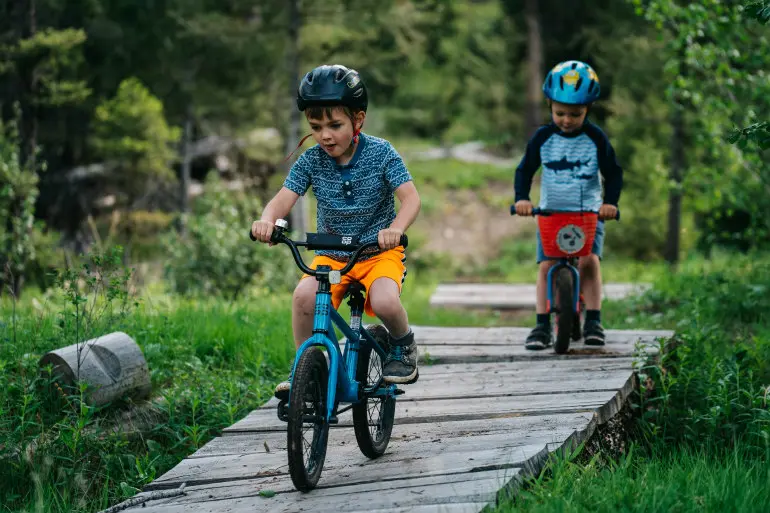 Two children ride bicycles on a wooden path through a wooded area.