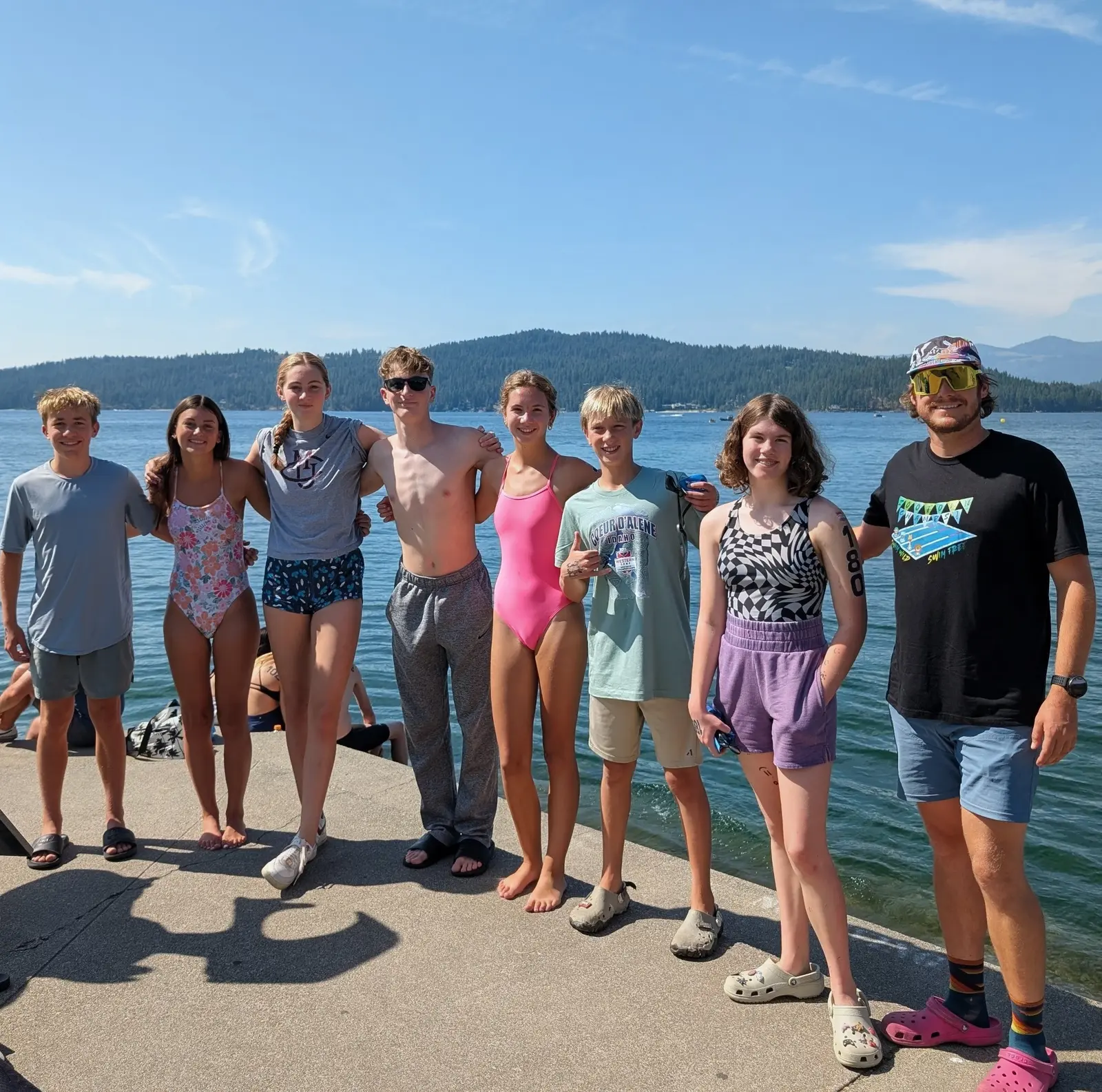 A group of young people and an adult instructor pose together on a dock near a lake.