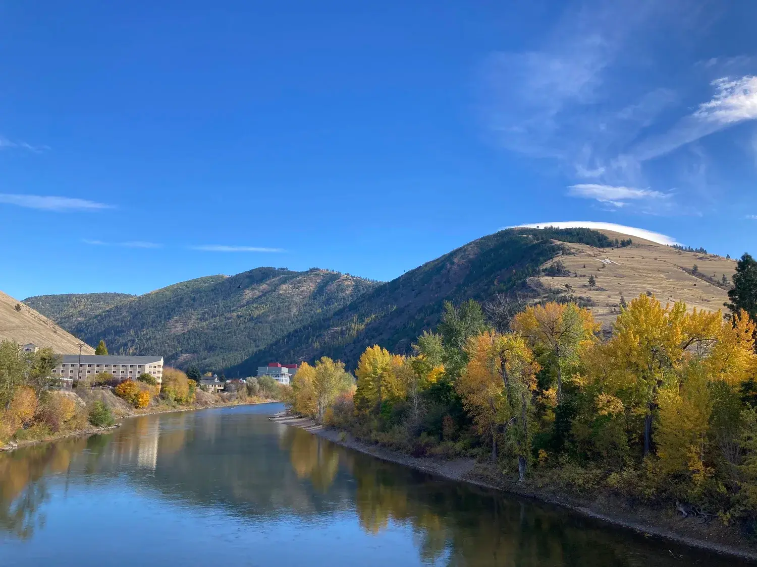 A river flows past golden autumn trees in front of a mountain landscape under a clear blue sky.