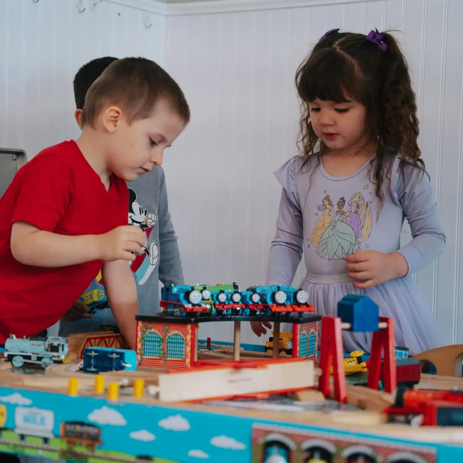 Three children are playing with toy trains on a wooden table.