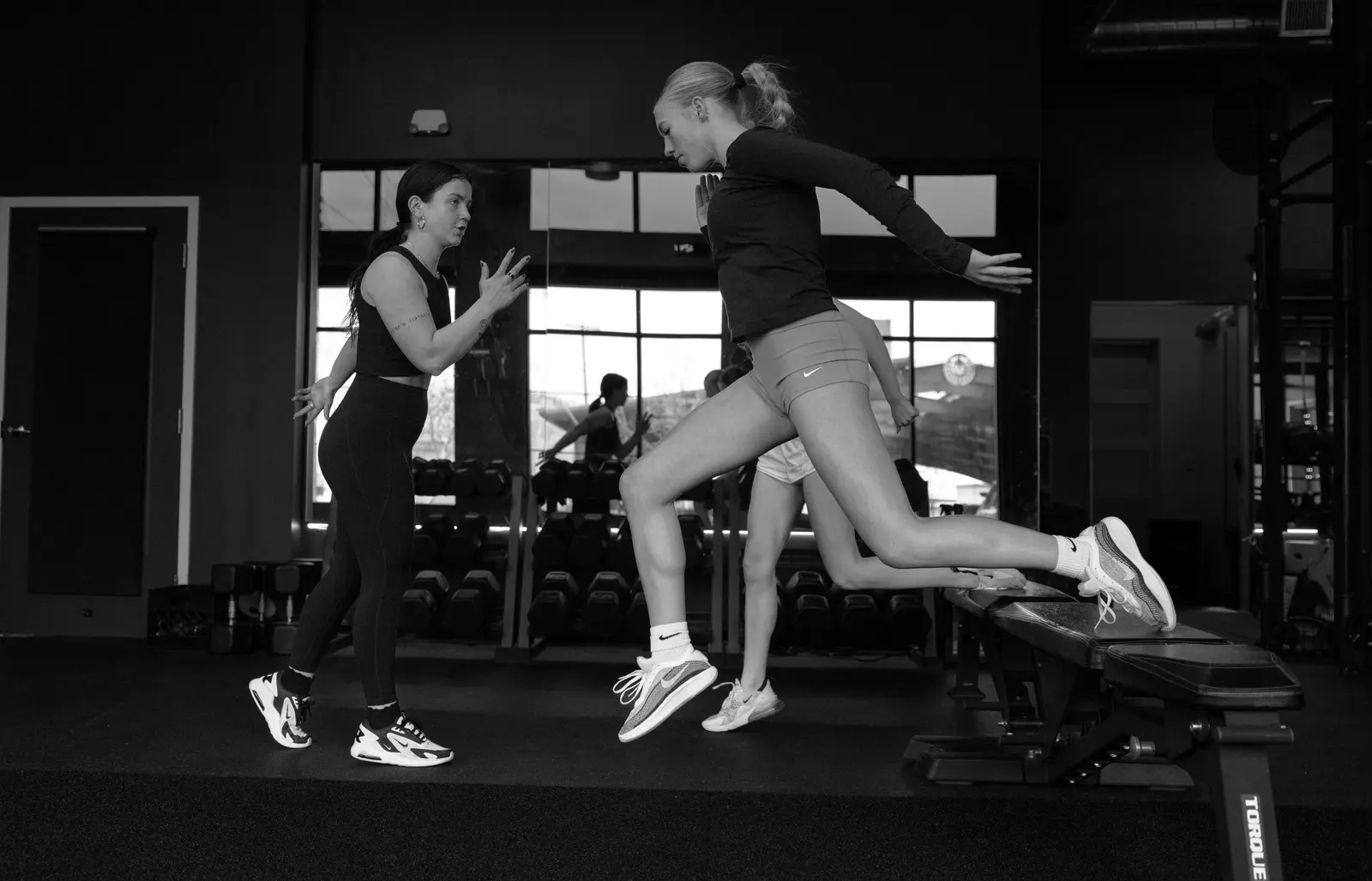 A fitness instructor coaches a woman performing a split squat on a bench in a gym.