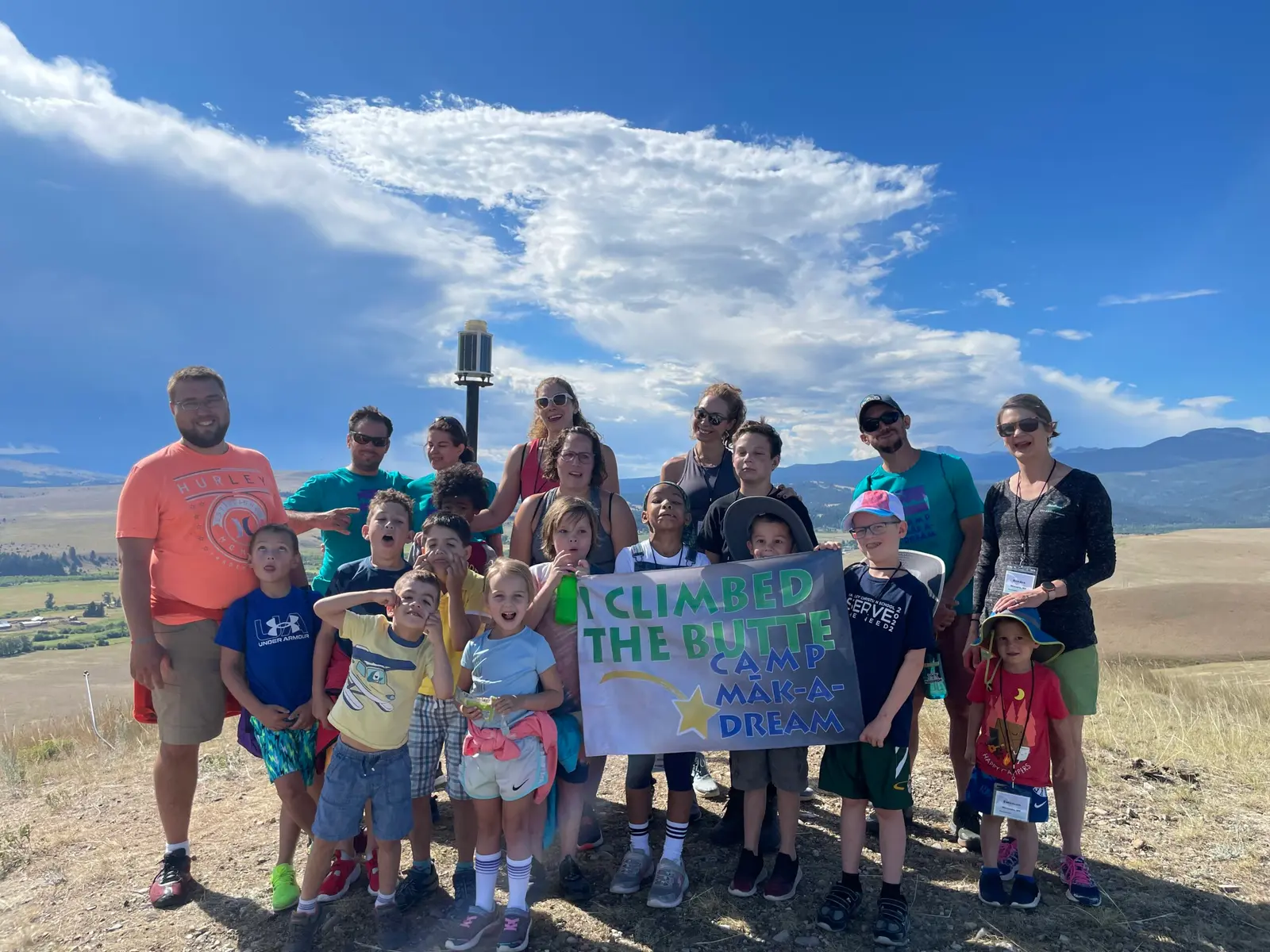 A large group of children and adults pose on a hilltop holding a banner.