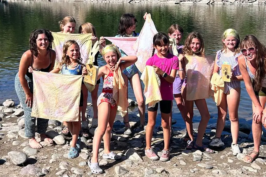 A group of children and two instructors pose by a river holding up pieces of dyed fabric.