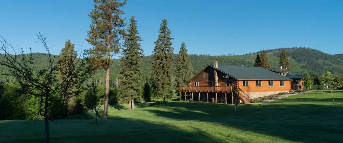 A large log cabin building sits in a grassy field surrounded by tall pine trees and a mountainous landscape.