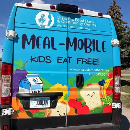 The rear view of a delivery van decorated with colorful food illustrations and text advertising a meal program for children.