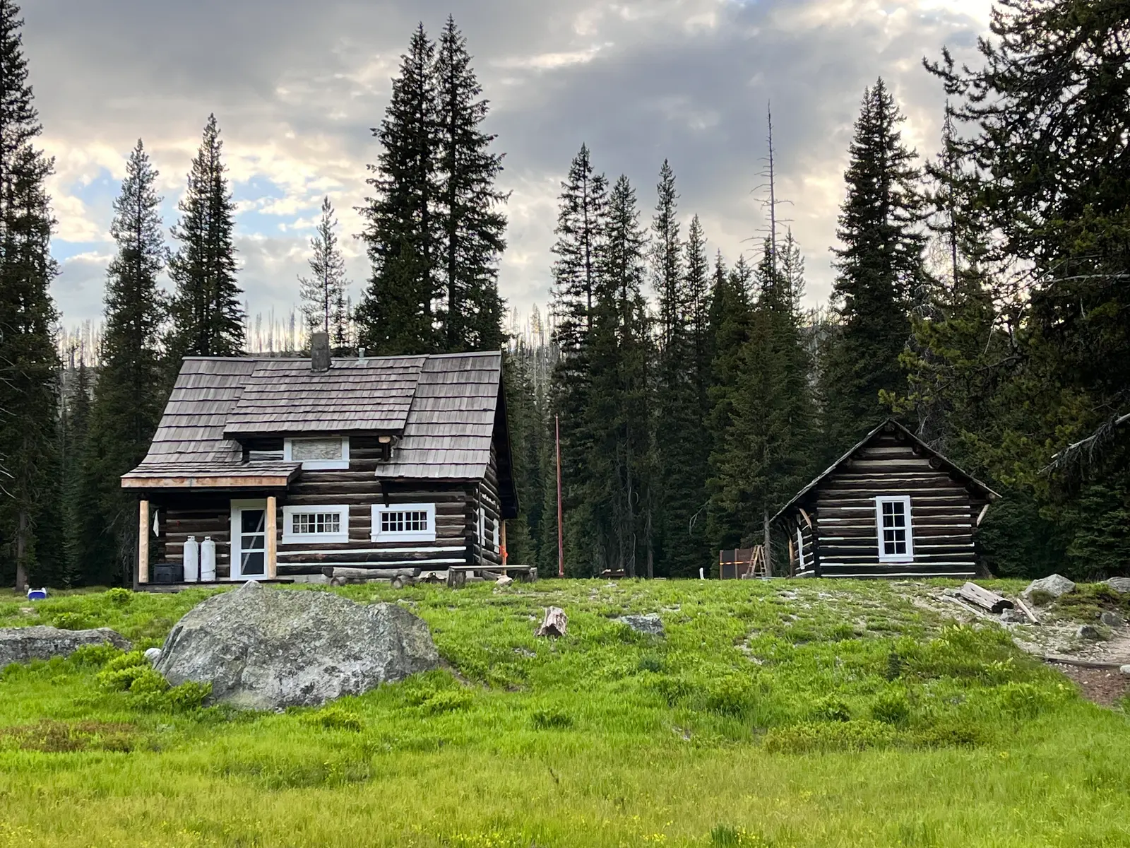 Two log cabins sit in a grassy meadow surrounded by a coniferous forest.