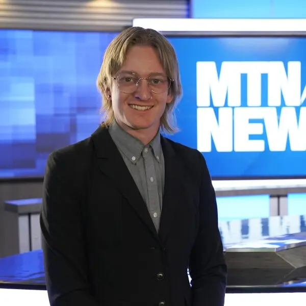 A person wearing a blazer and glasses stands in a news studio.