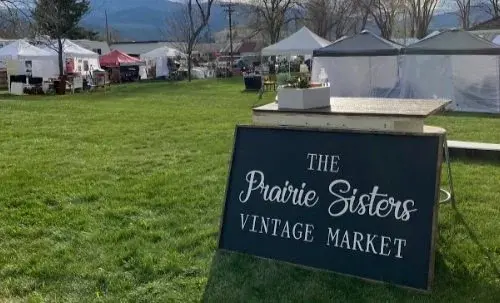 A sign for a vintage market stands in a grassy field with event tents in the background.