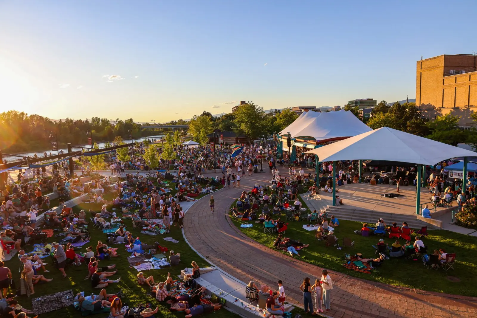 A large crowd of people gather on a lawn near a riverside pavilion during an outdoor evening event.