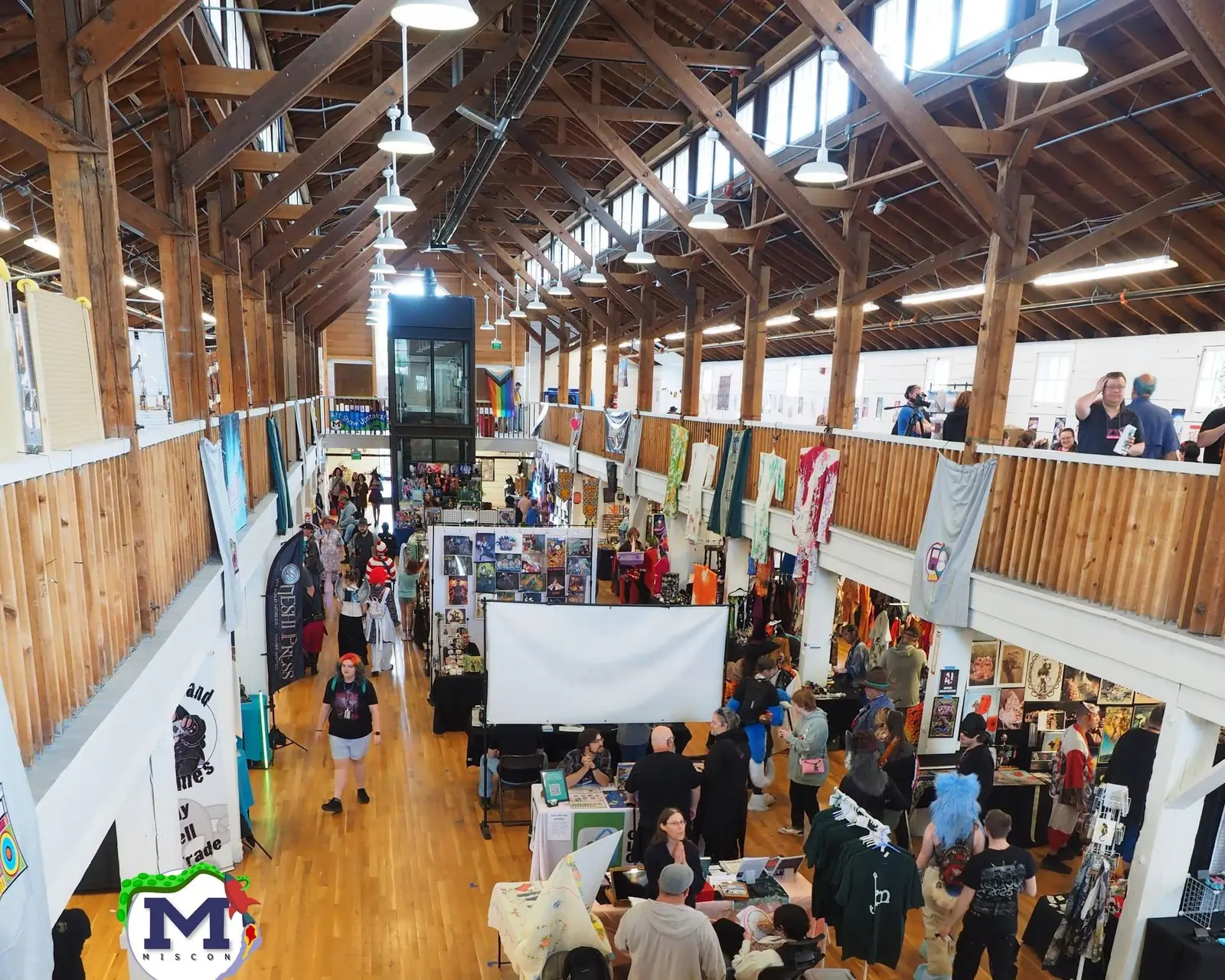 A high-angle view shows people browsing vendors in a large wooden hall at an event.