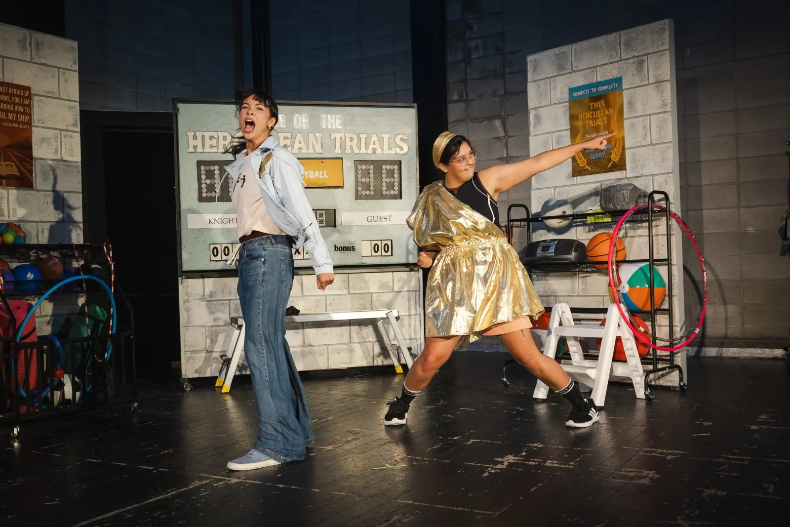 Two performers in costume act on a stage with a gymnasium set.