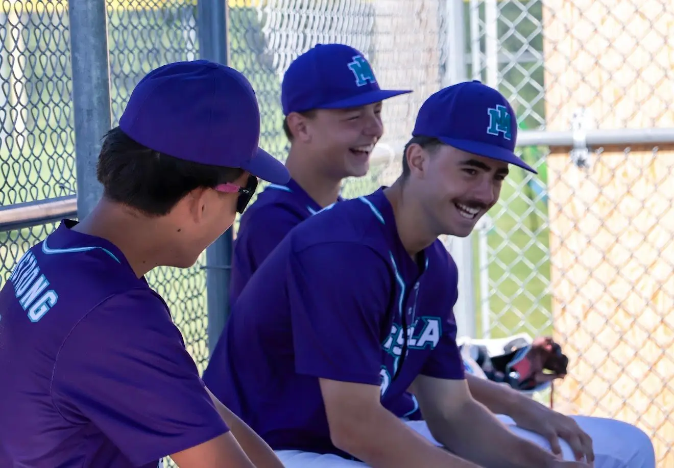 Three baseball players in purple uniforms sit on a bench and laugh.