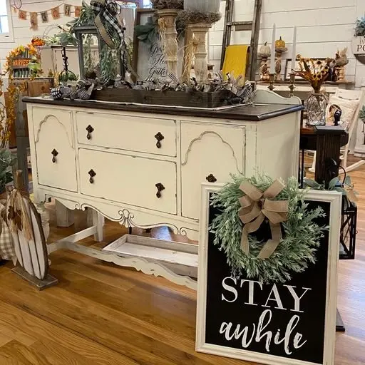 Decorative display table with a wreath and a sign saying "Stay Awhile" in a store with various home decor items.