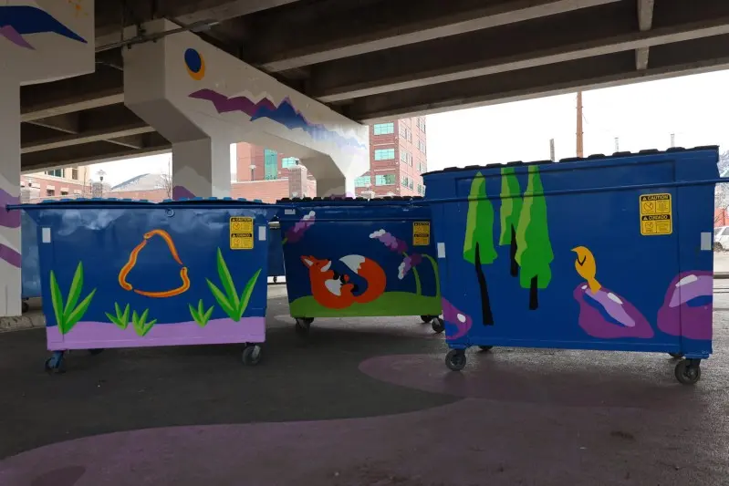 Three blue dumpsters with painted animal and nature scenes sit under a bridge.