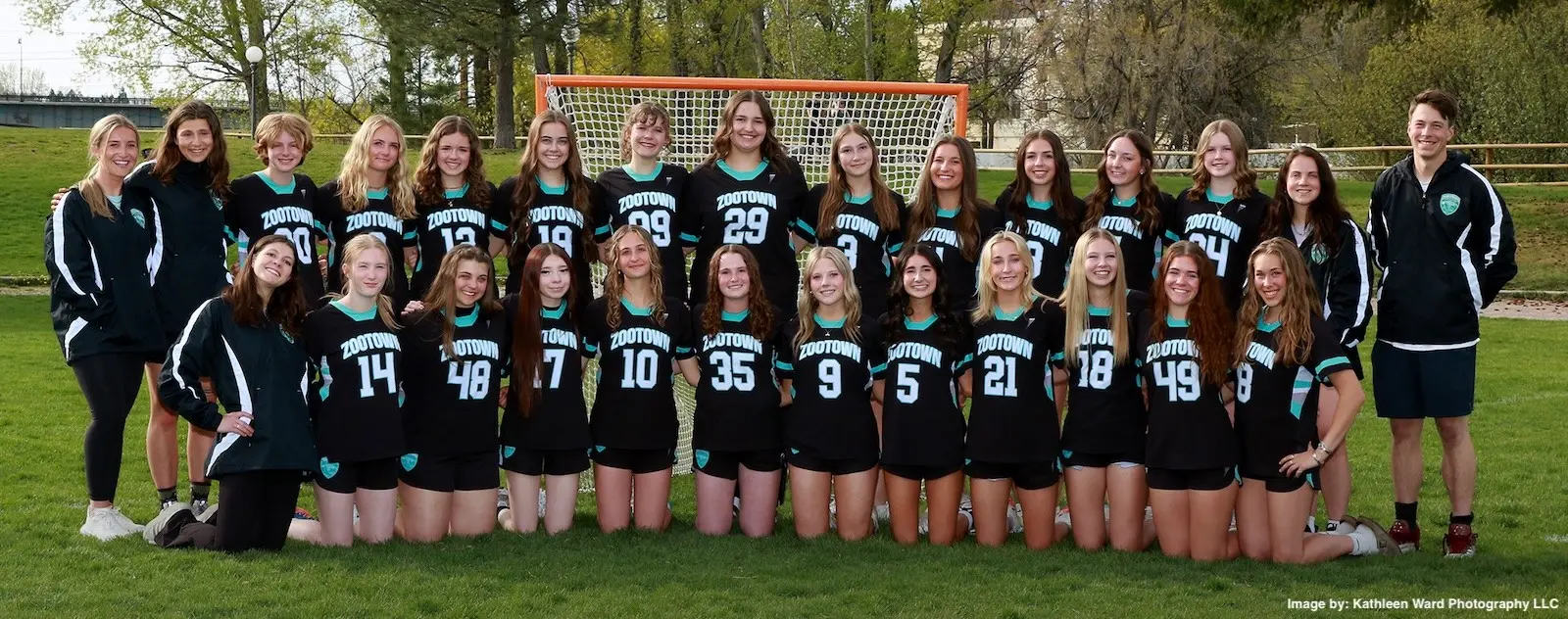 A large group of female athletes in black and teal jerseys pose in front of a lacrosse goal with their coach.