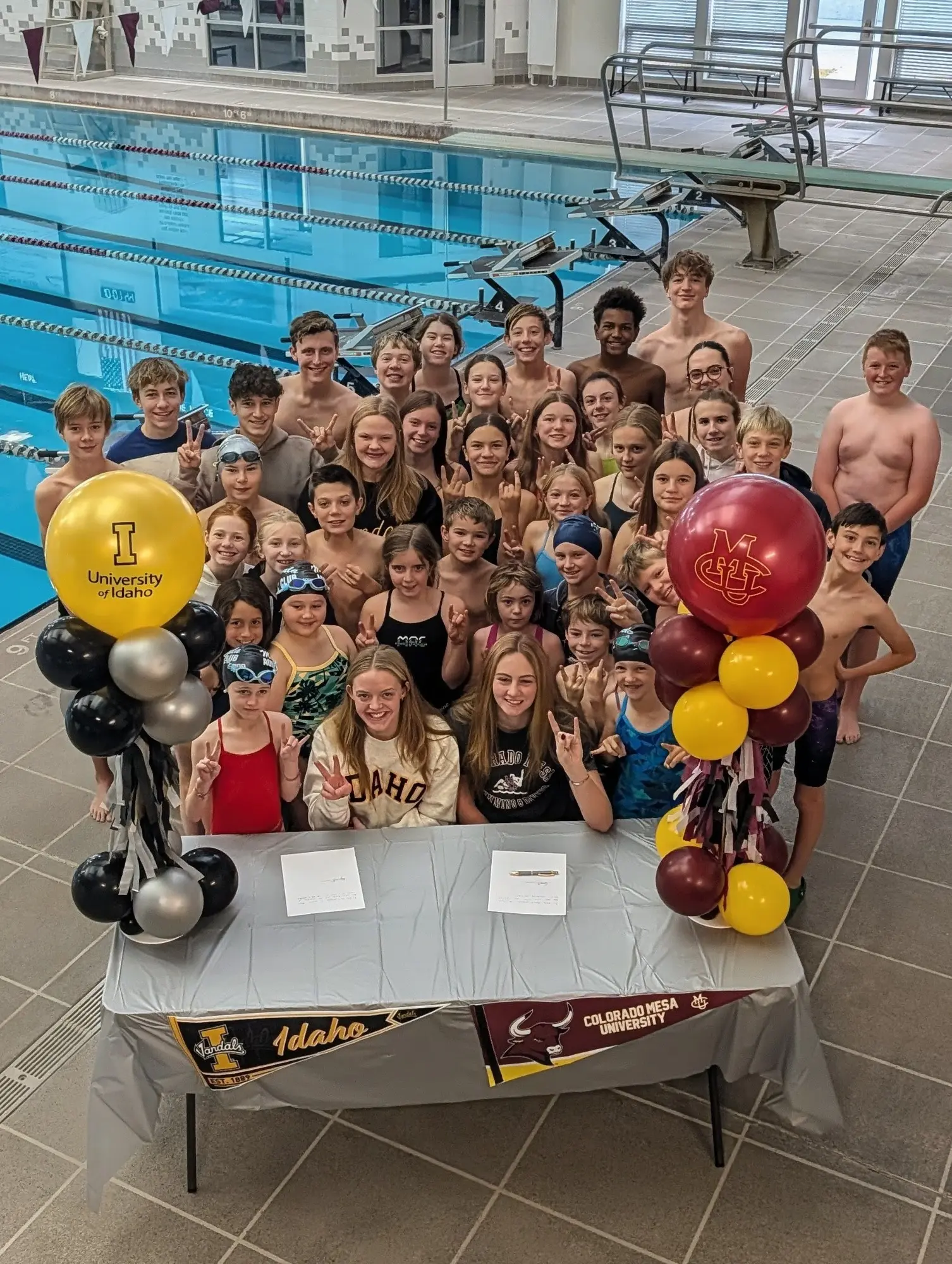 A large group of swimmers poses behind a signing table at a pool.