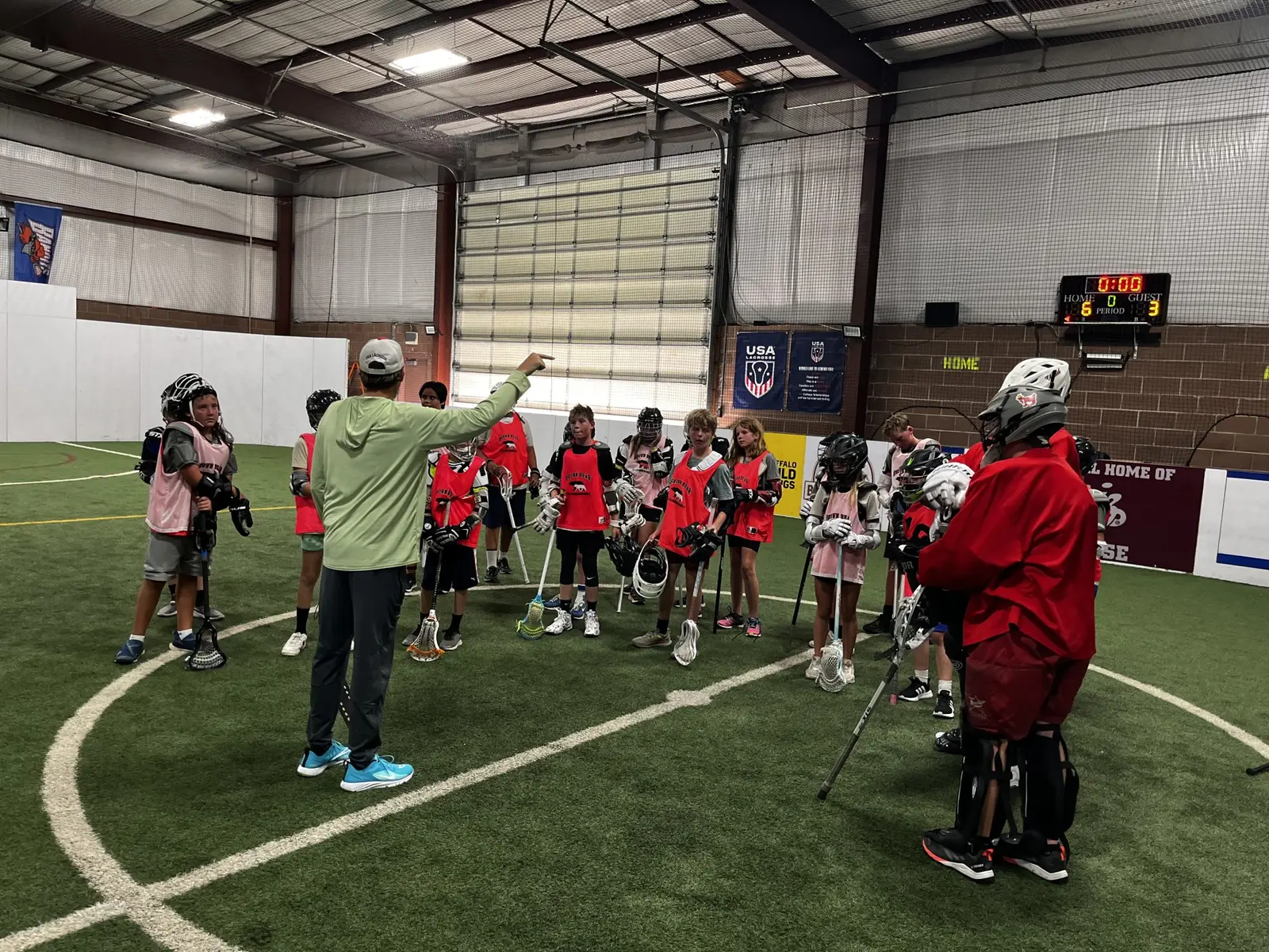A coach points while talking to a group of lacrosse players on an indoor field.