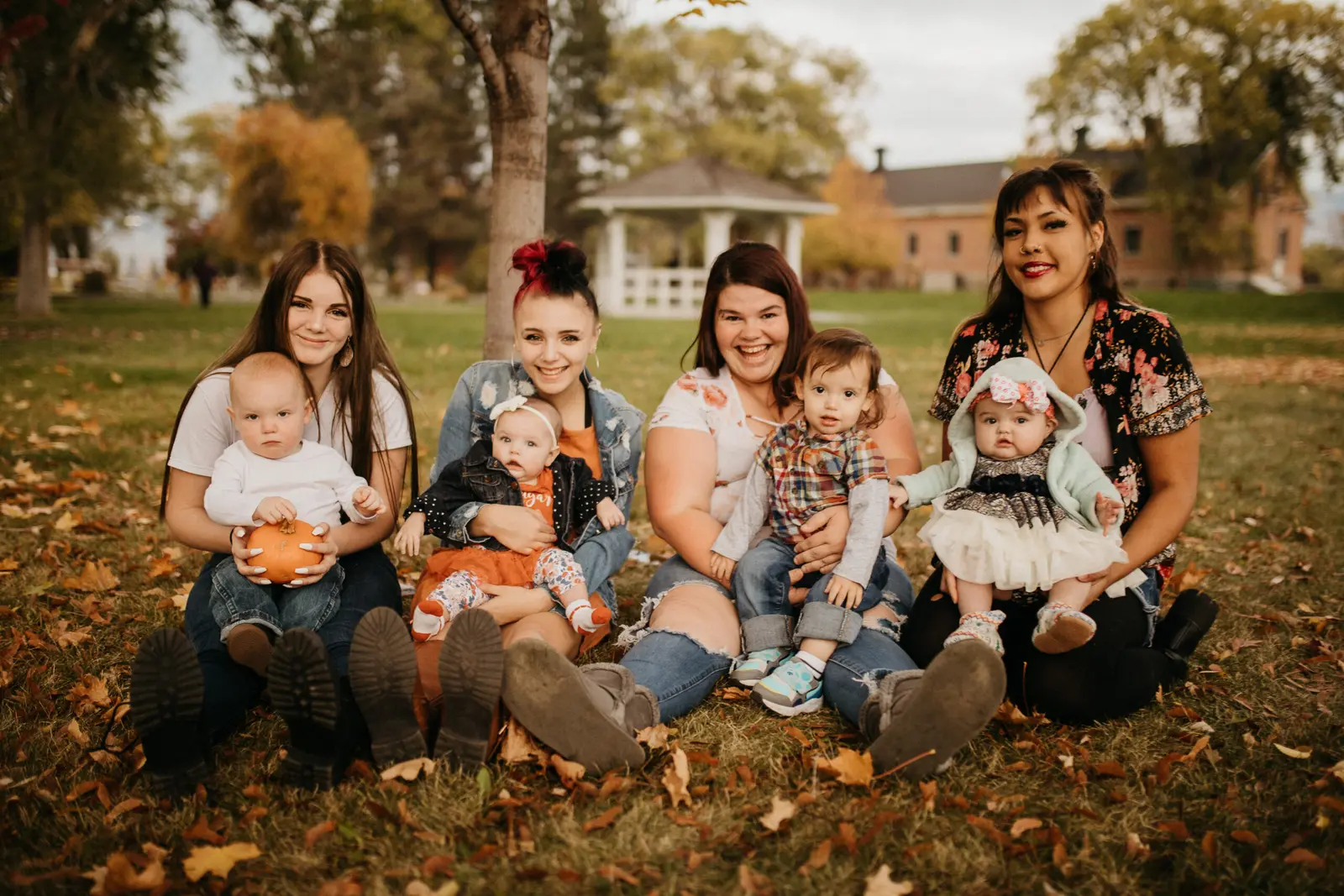 Four women are sitting on the grass with their infants in an outdoor park setting.