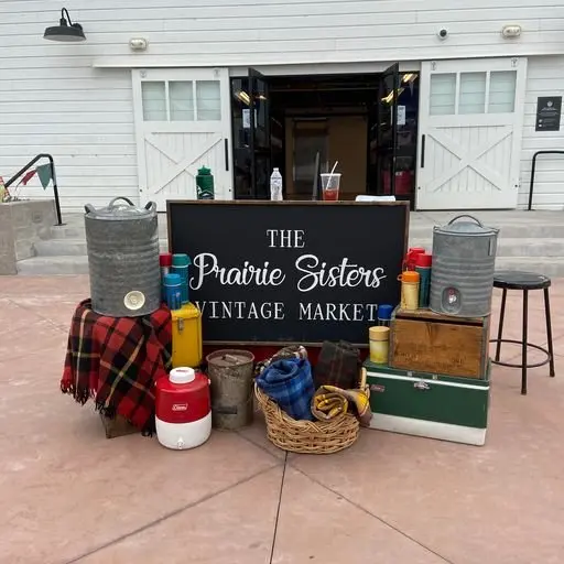 Outdoor vintage market display with a blackboard sign reading 'The Prairie Sisters Vintage Market' surrounded by vintage items like coolers, blankets, baskets, and decorative cans in front of a white building.