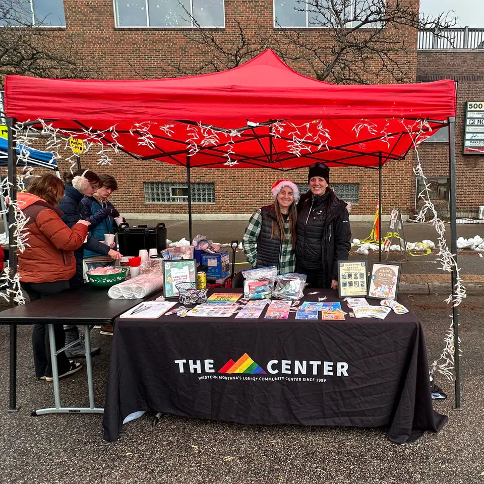 Two women stand at a booth for a community center under a red canopy in a parking lot.