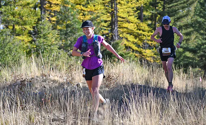 A woman and a man run along a grassy outdoor trail.
