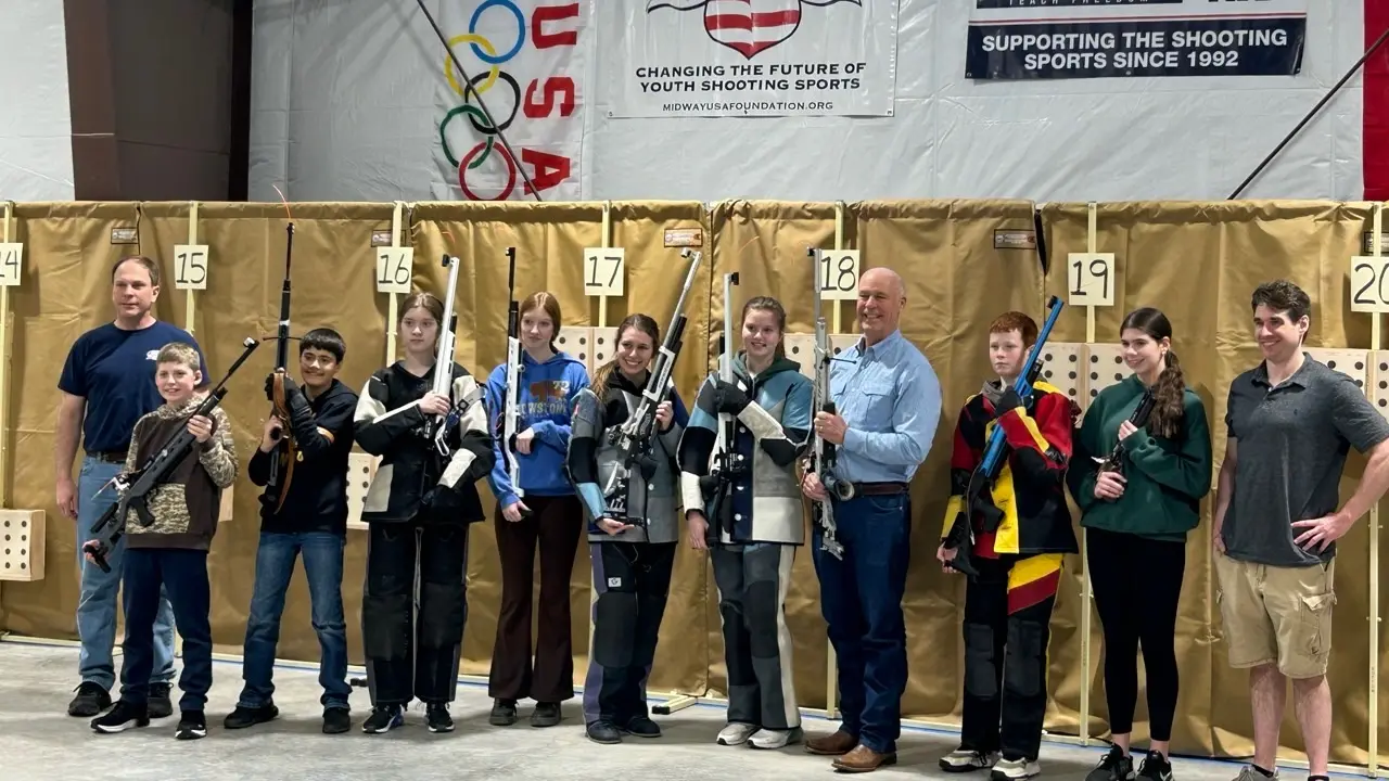 A group of youth and adults pose with their rifles at a shooting sports event.