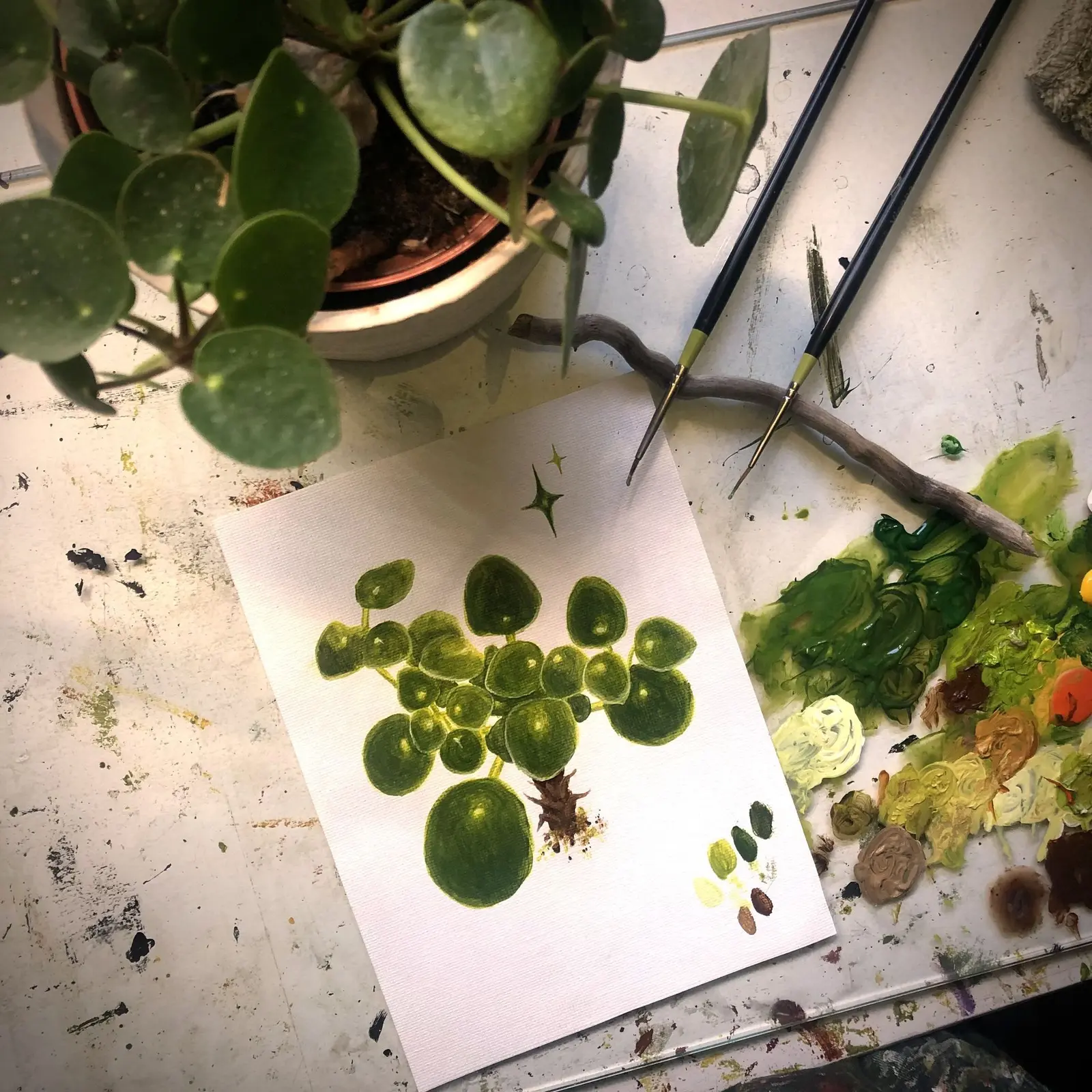 A painting of a plant sits on a table next to brushes, paint, and the actual plant it depicts.