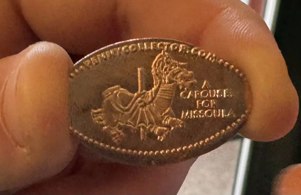 A hand holds an elongated pressed penny souvenir coin featuring a carousel horse design.