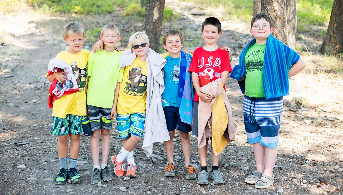 Six young boys stand together in a wooded area wearing casual summer clothes and carrying towels.