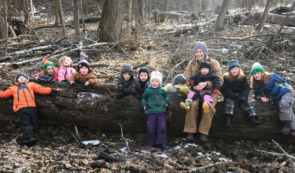 A group of children and an instructor are sitting on a large fallen log in a wooded area.