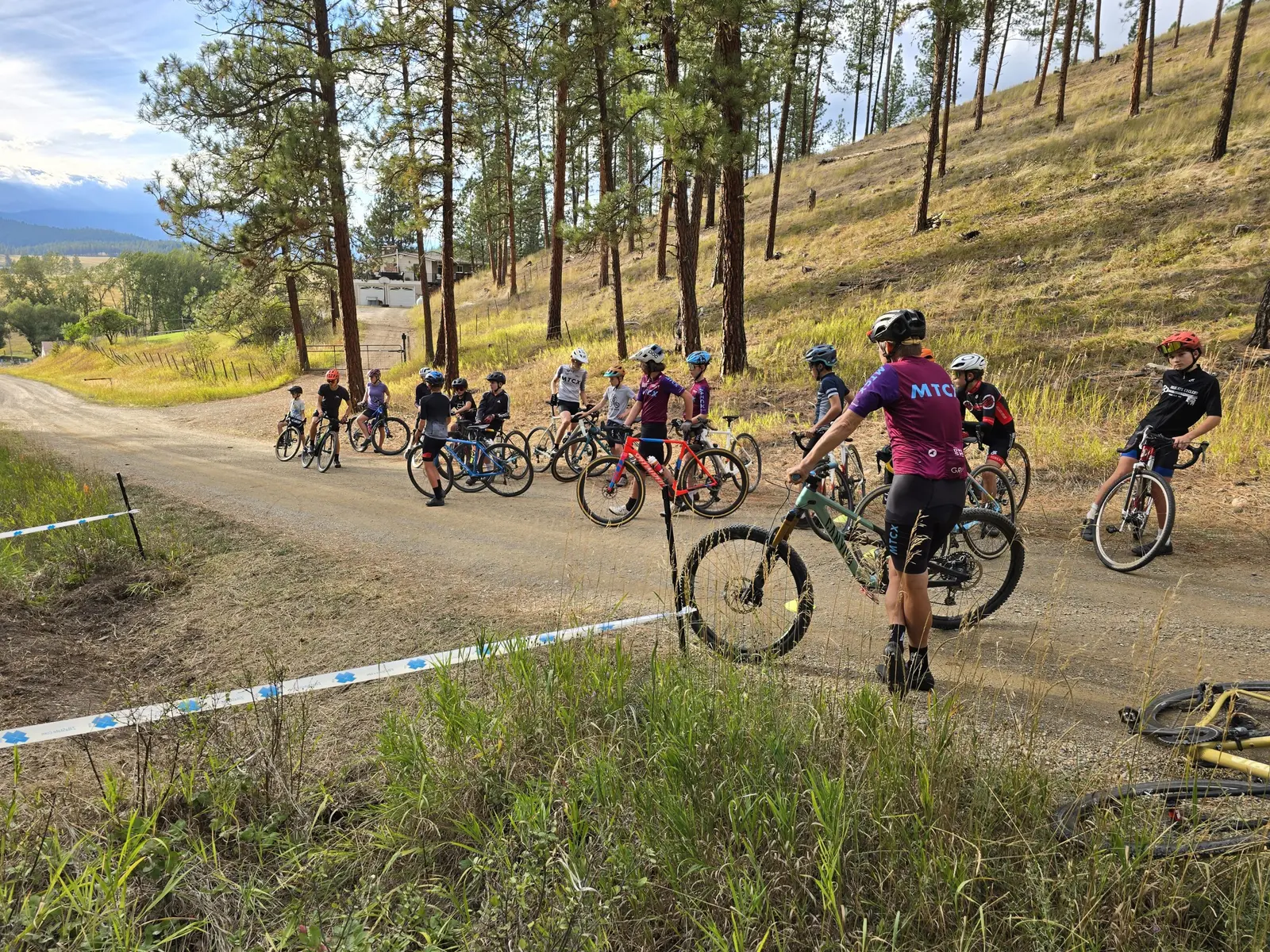 A group of young cyclists wearing helmets gather on a gravel road near a forest.