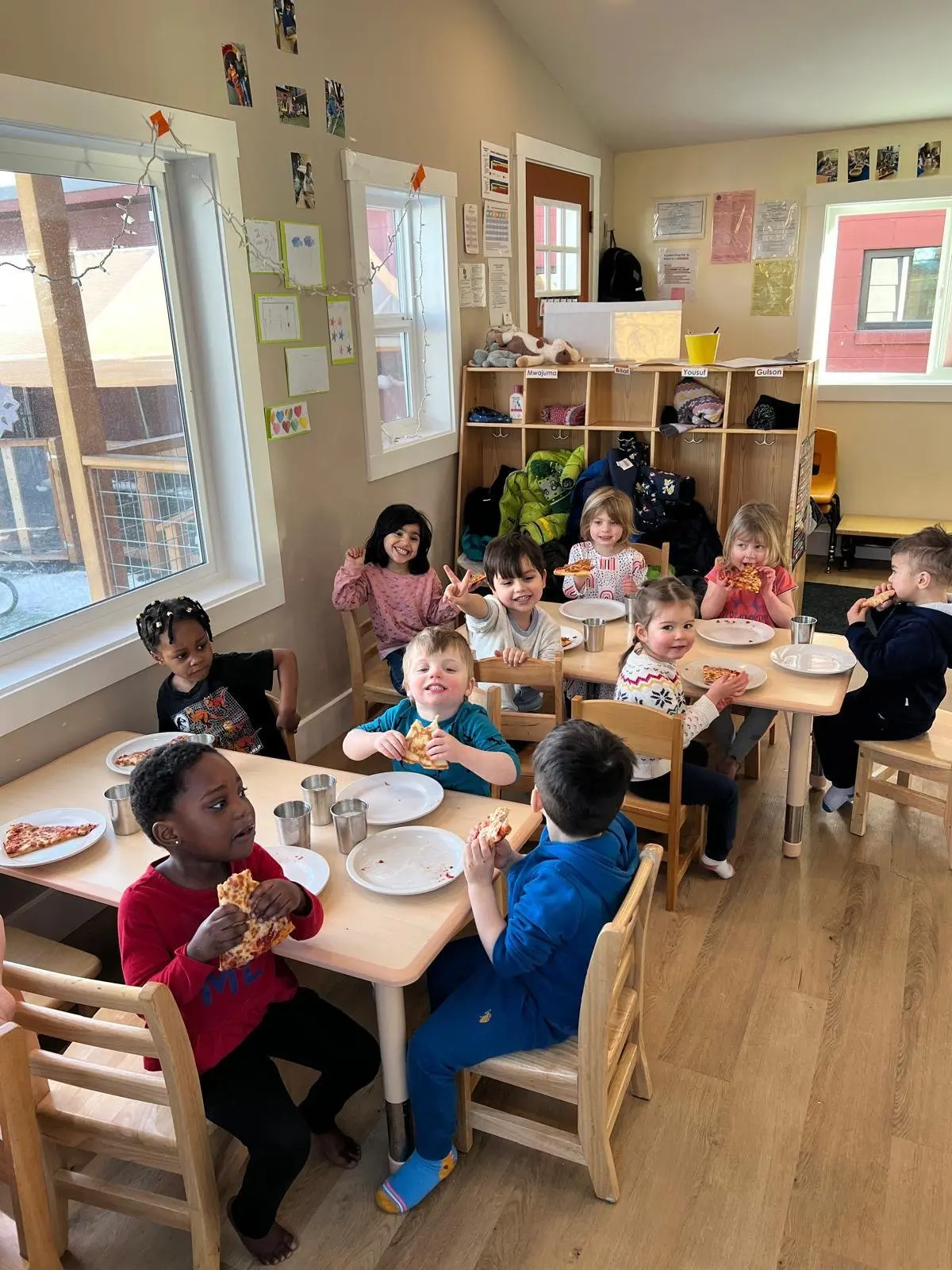 Children sitting at tables in a classroom eating pizza and smiling.