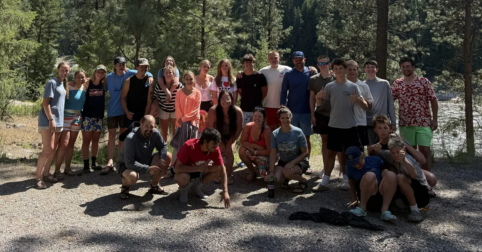 A large group of young adults and teenagers poses for a photo in a wooded, riverside area.