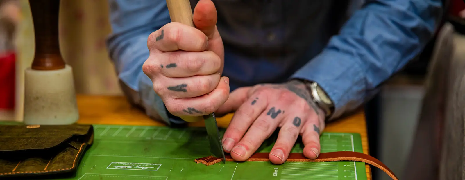 A person's tattooed hands use a tool to cut a strip of leather on a green cutting mat.