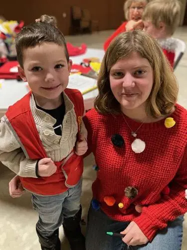 A young boy and a woman wearing a red sweater with pom-poms pose together for a photo.