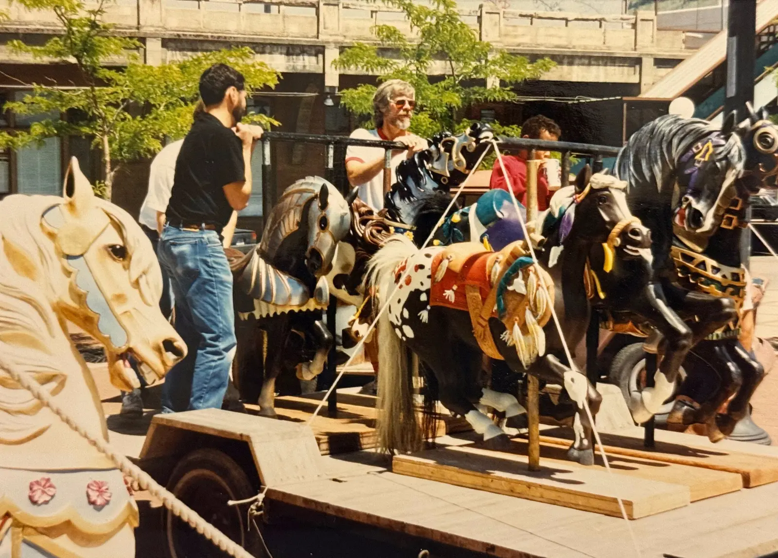 Men stand beside a carousel of painted horses on a portable trailer outdoors.