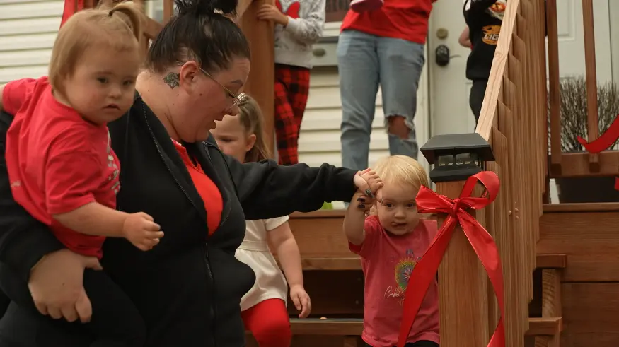 A caregiver holds a young child while another child stands nearby on a wooden staircase decorated with a red ribbon.