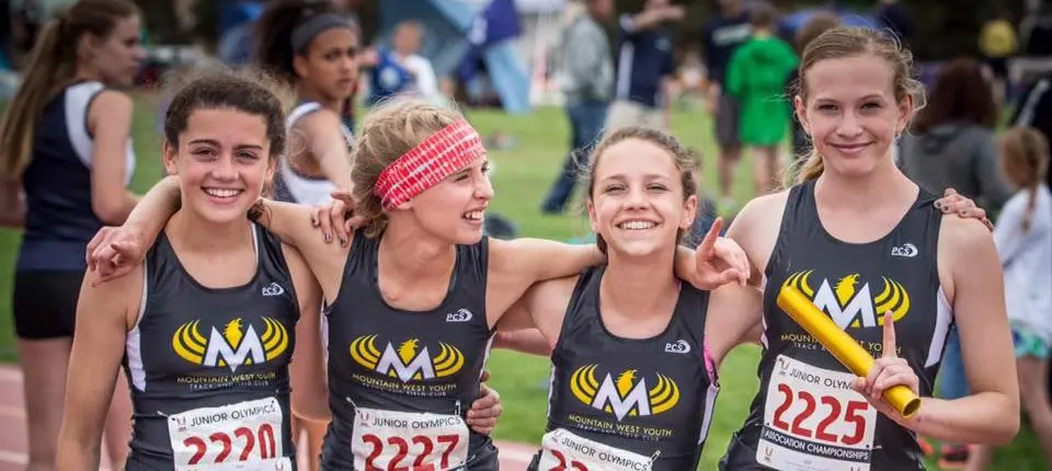 Four young female track athletes wearing matching uniforms smile together on a field.