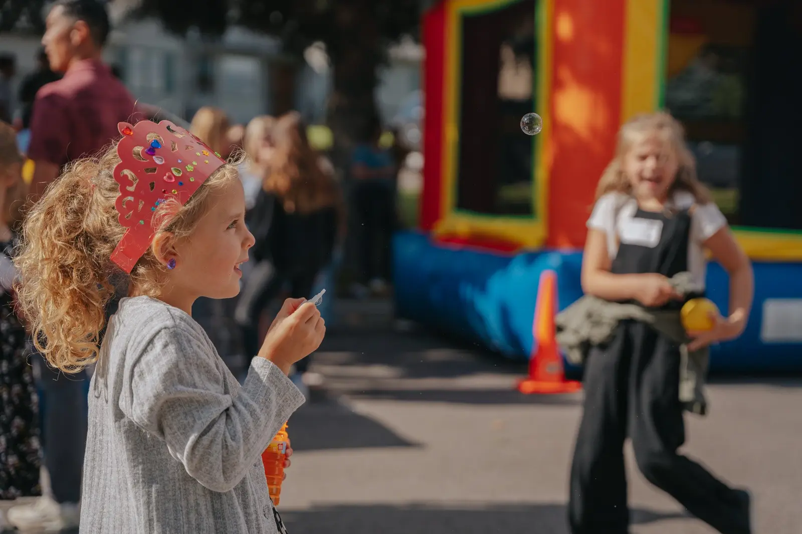 A young girl wearing a crown plays with bubbles at an outdoor event.