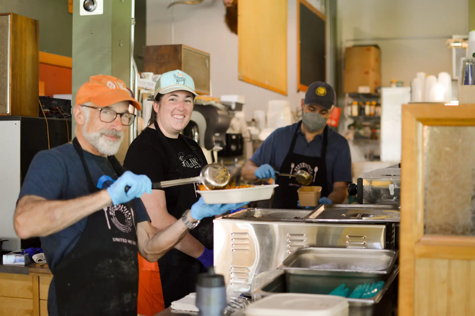 Three people wearing aprons and gloves work in a kitchen preparing food for service.
