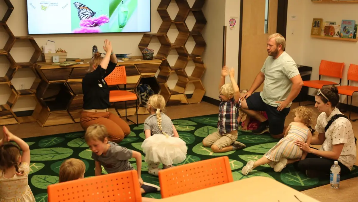 An instructor leads a group of children and adults in an activity on a leaf-patterned rug.