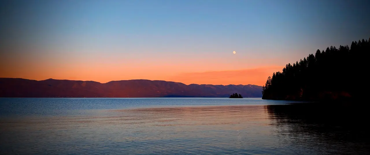 The sun sets over a calm lake with mountains in the distance and a moon visible in the sky.