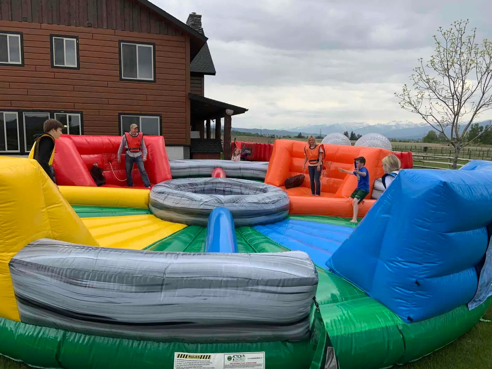 Children and adults playing on a large, colorful inflatable water bounce house outside in a backyard with a house, trees, and mountains in the distance.
