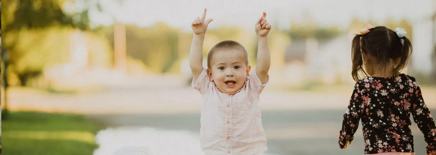A young boy with his arms raised stands next to a girl in a floral shirt outdoors.
