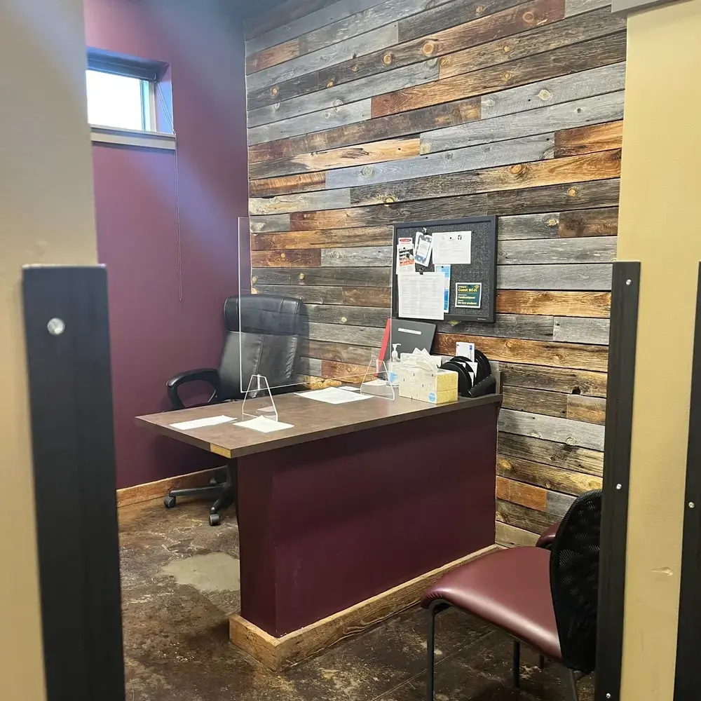 An office workspace features a desk with a protective screen and a reclaimed wood accent wall.