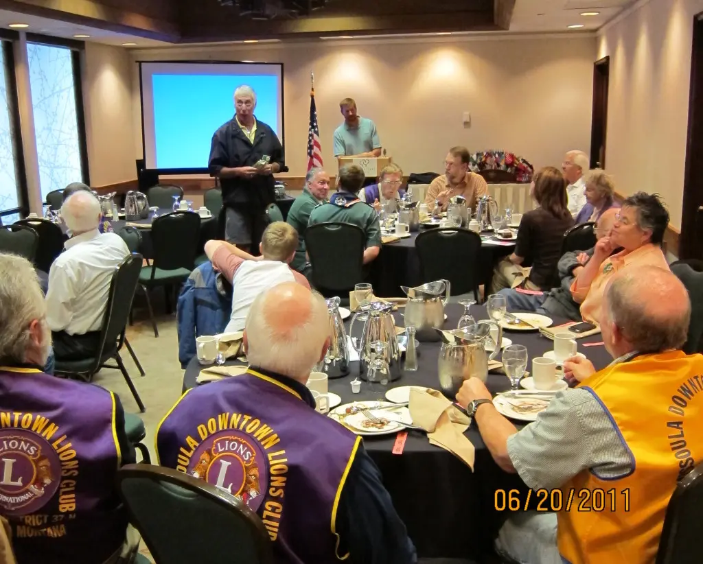 A group of people gather in a room for a luncheon or meeting, with some attendees wearing club vests.