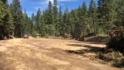 Photo from nbcmontana.com Construction equipment operates on a cleared dirt lot bordered by a dense evergreen forest.