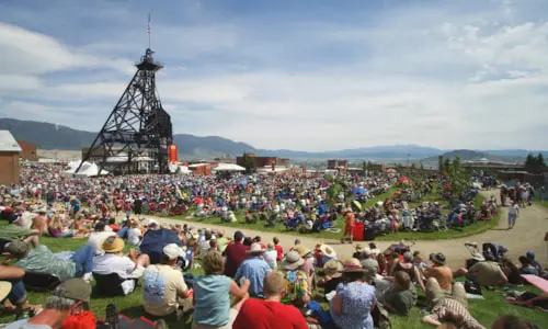 A large crowd of people sits on a grassy hillside watching an outdoor event near a metal tower structure.